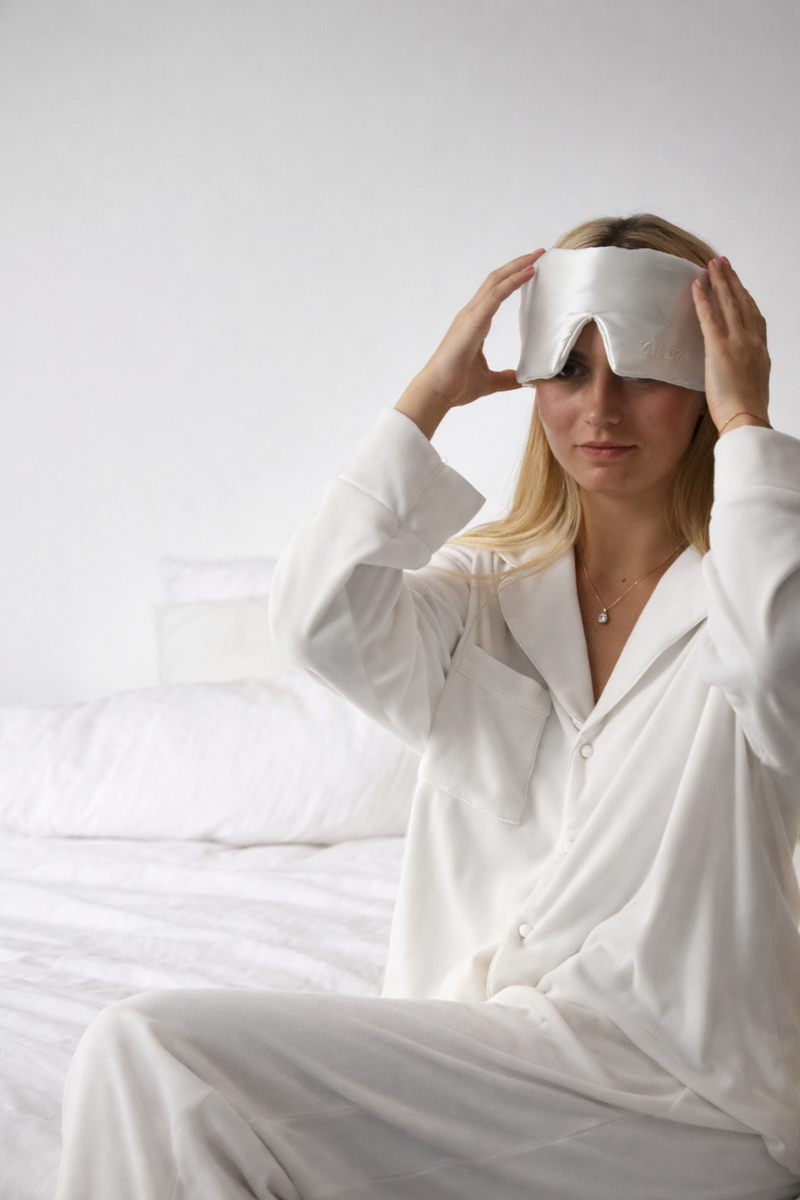 Woman in white sleepwear putting on a white Still Suain silk sleep mask